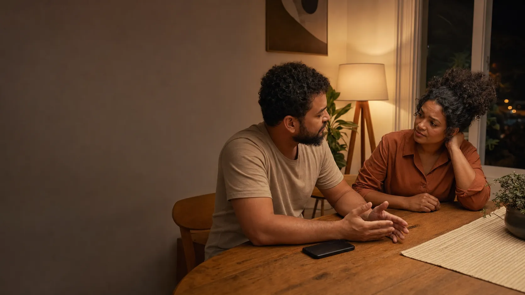 Dois adultos conversam com atenção em uma mesa de casa, em ambiente acolhedor.